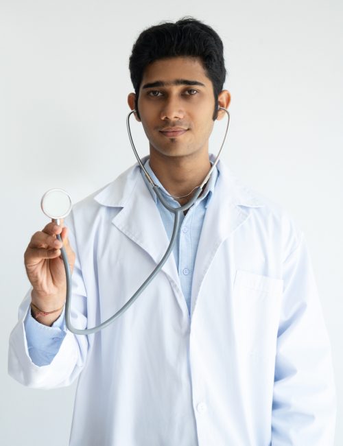 Serious male general practitioner using stethoscope Serious male general practitioner using stethoscope and looking at camera. Concentrated young Indian doctor in lab coat holding medical examination and listening to lung sounds. Medical device concept