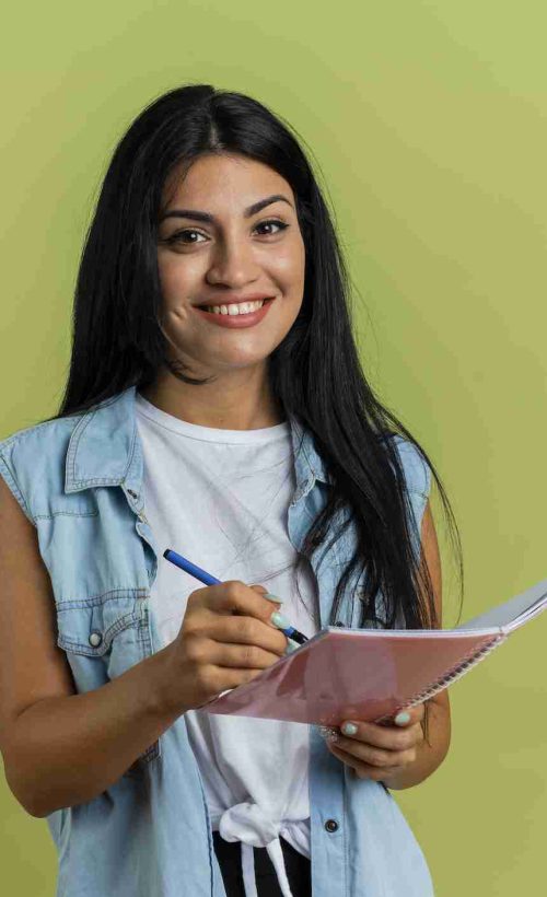smiling young caucasian girl holds pen and notebook looking at camera isolated on olive green background with copy space